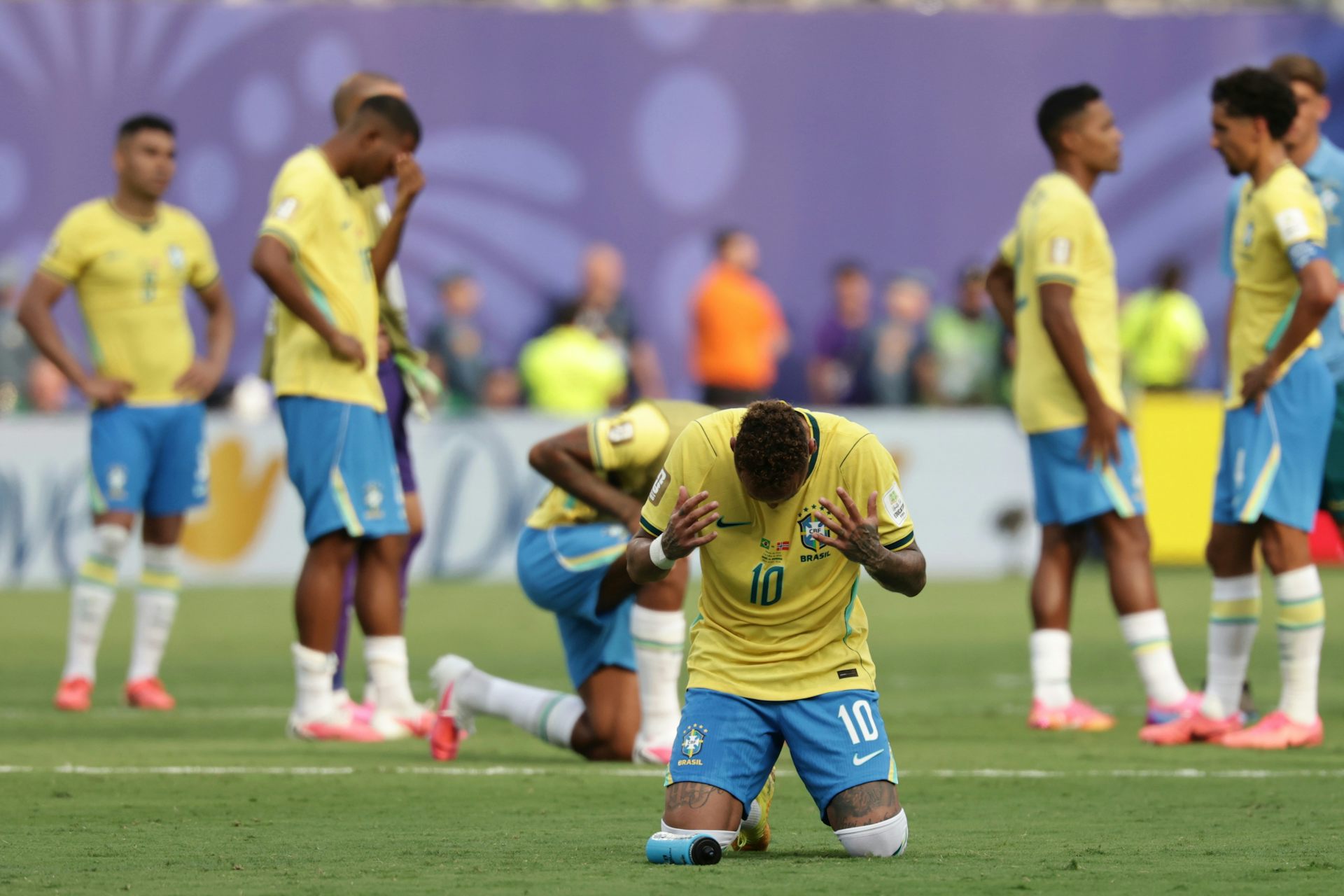un homme vêtu d'un t-shirt jaune et d'un short bleu est à genoux sur un terrain de foot, les mains jointes en signe de prière ; d'autres hommes déambulent derrière lui