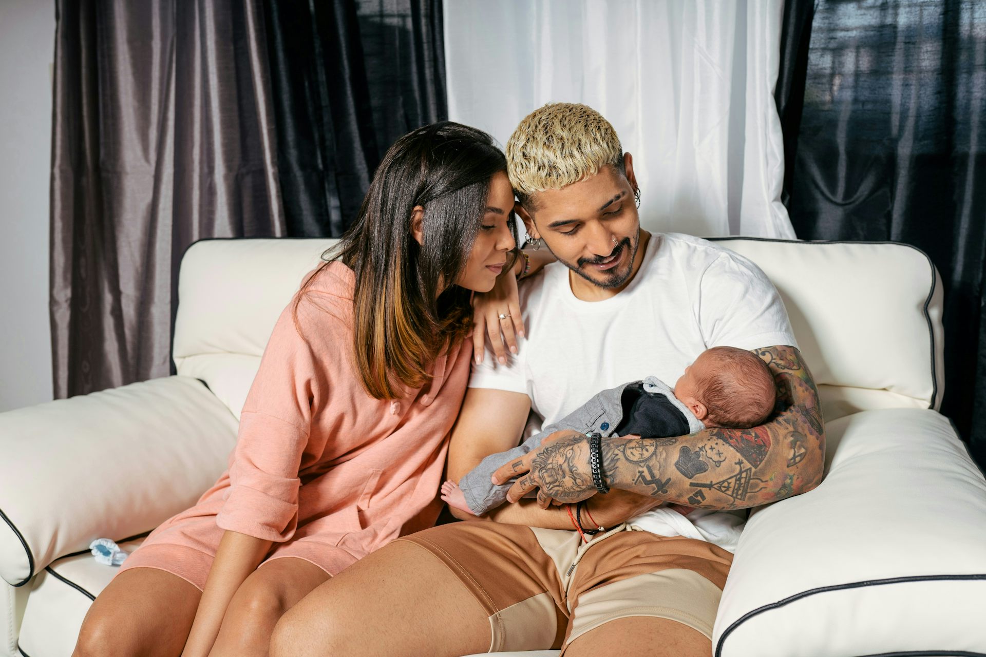Deux jeunes parents (femme et homme) sont assis près l'un de l'autre sur un canapé, et le père tient le bébé dans ses bras.