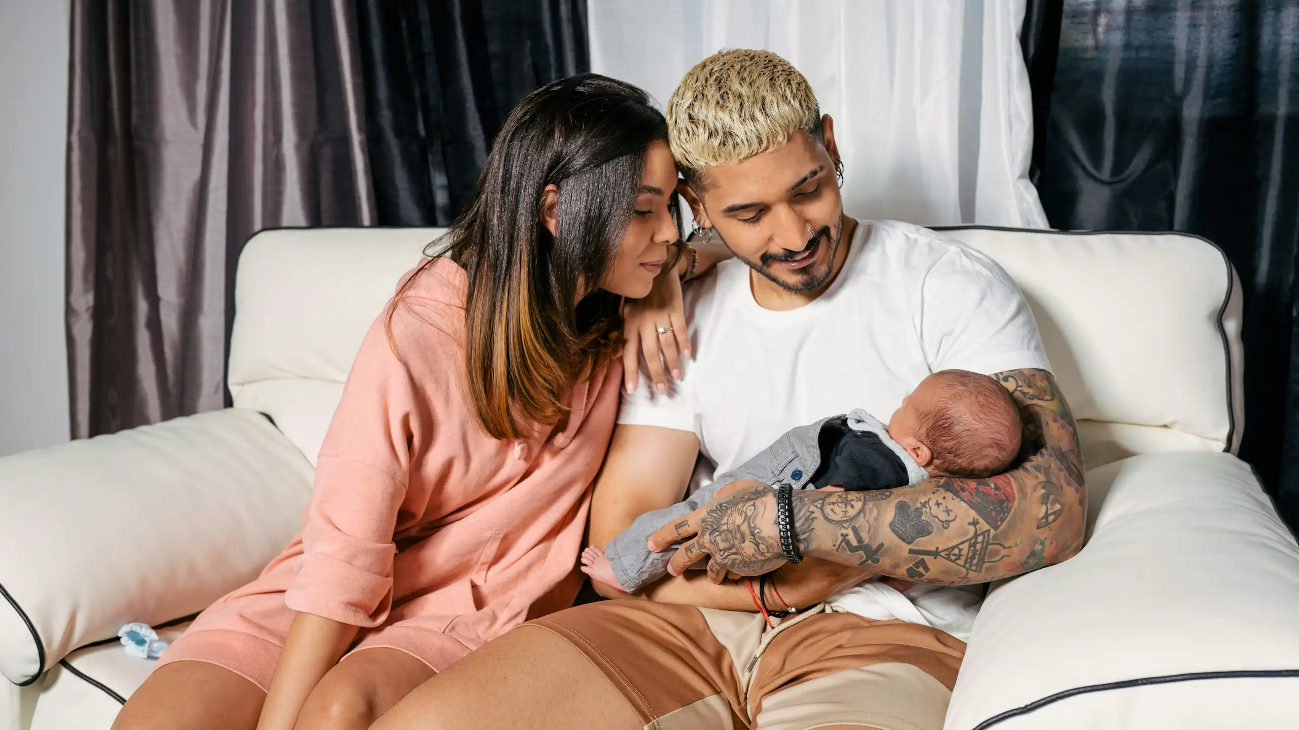Deux jeunes parents (femme et homme) sont assis près l'un de l'autre sur un canapé, et le père tient le bébé dans ses bras.