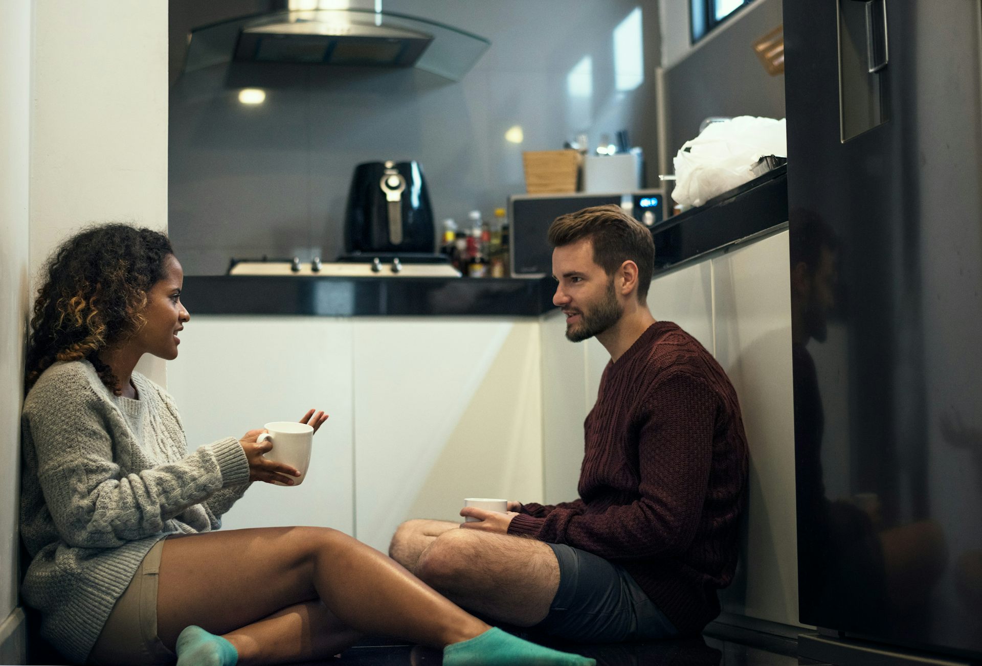 Une jeune femme et un jeune homme) discutent ensemble, assis sur le plancher d’une cuisine