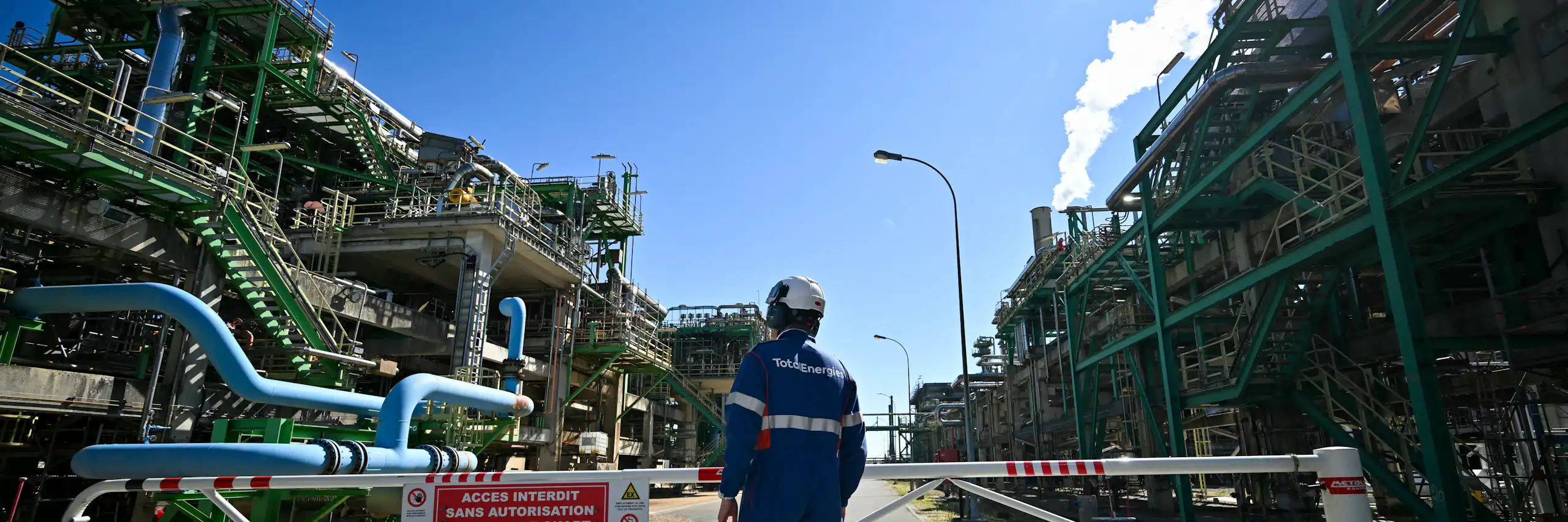 A person in a helmet and protective equipment stands on a paved area between two massive industrial buildings covered with tubes, wires and staircases.