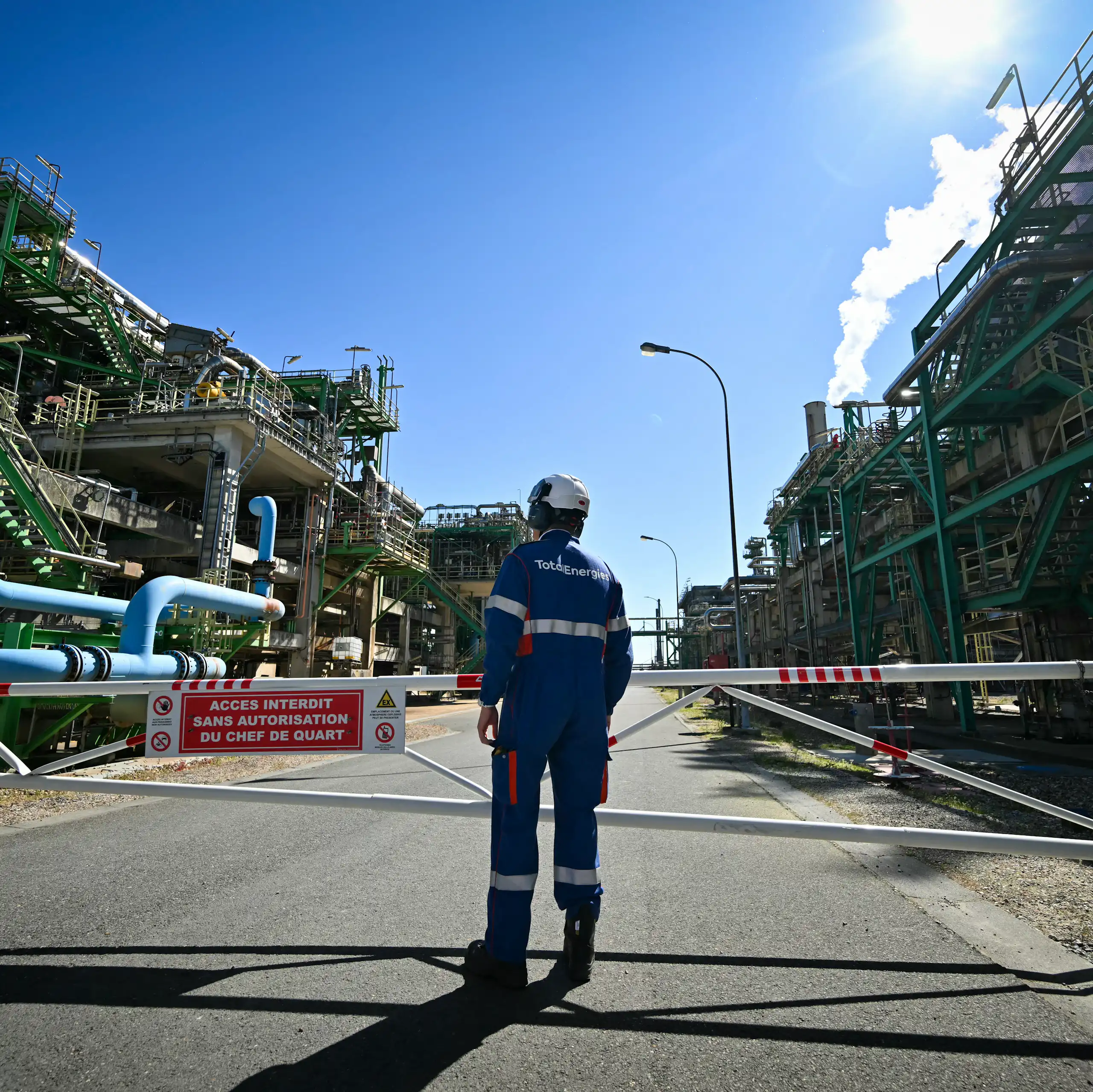 A person in a helmet and protective equipment stands on a paved area between two massive industrial buildings covered with tubes, wires and staircases.