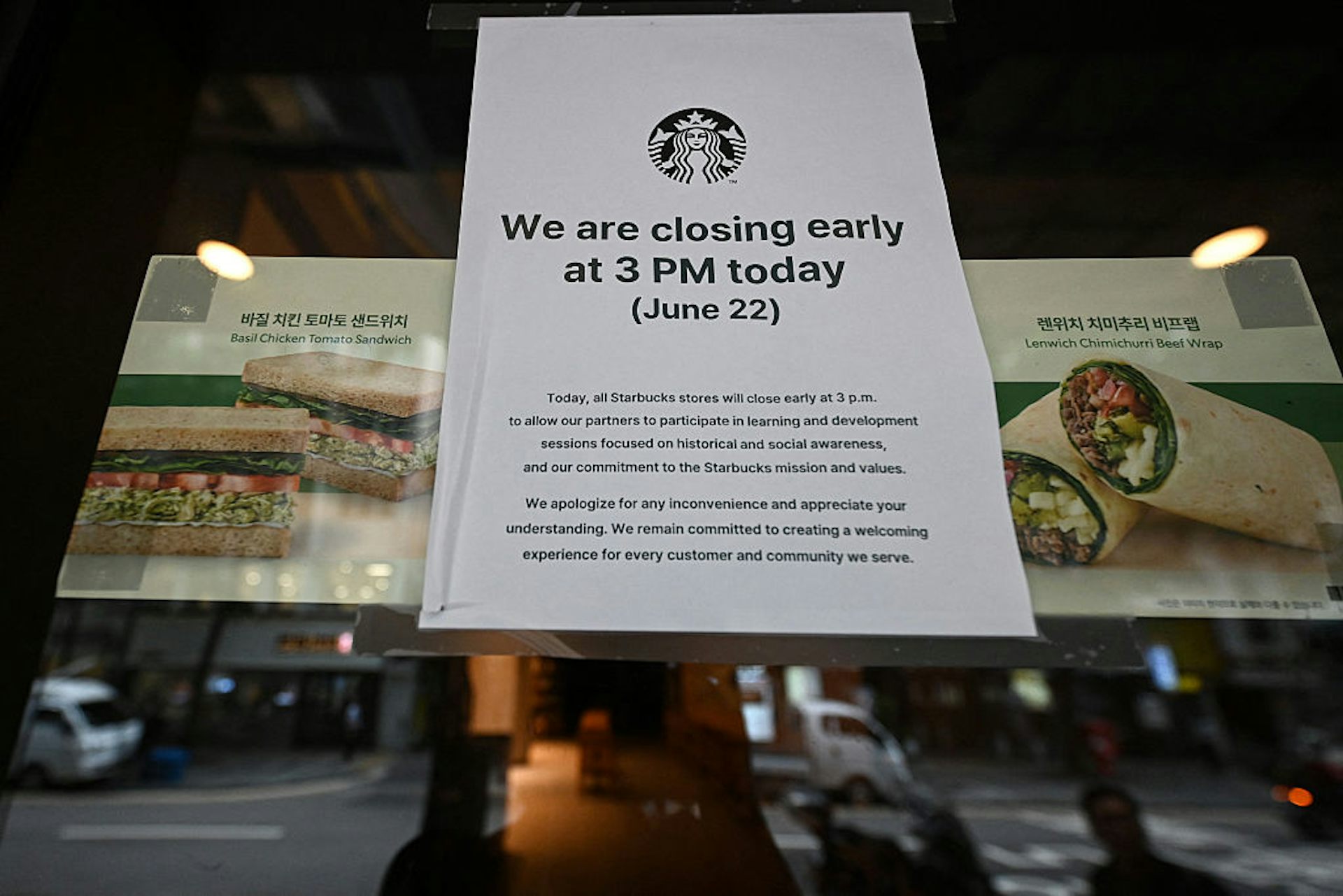 A glass window of a Starbucks location featuring posters of sandwiches and wraps alongside a white sign announcing an early closing so staff could undergo historical and social awareness training.