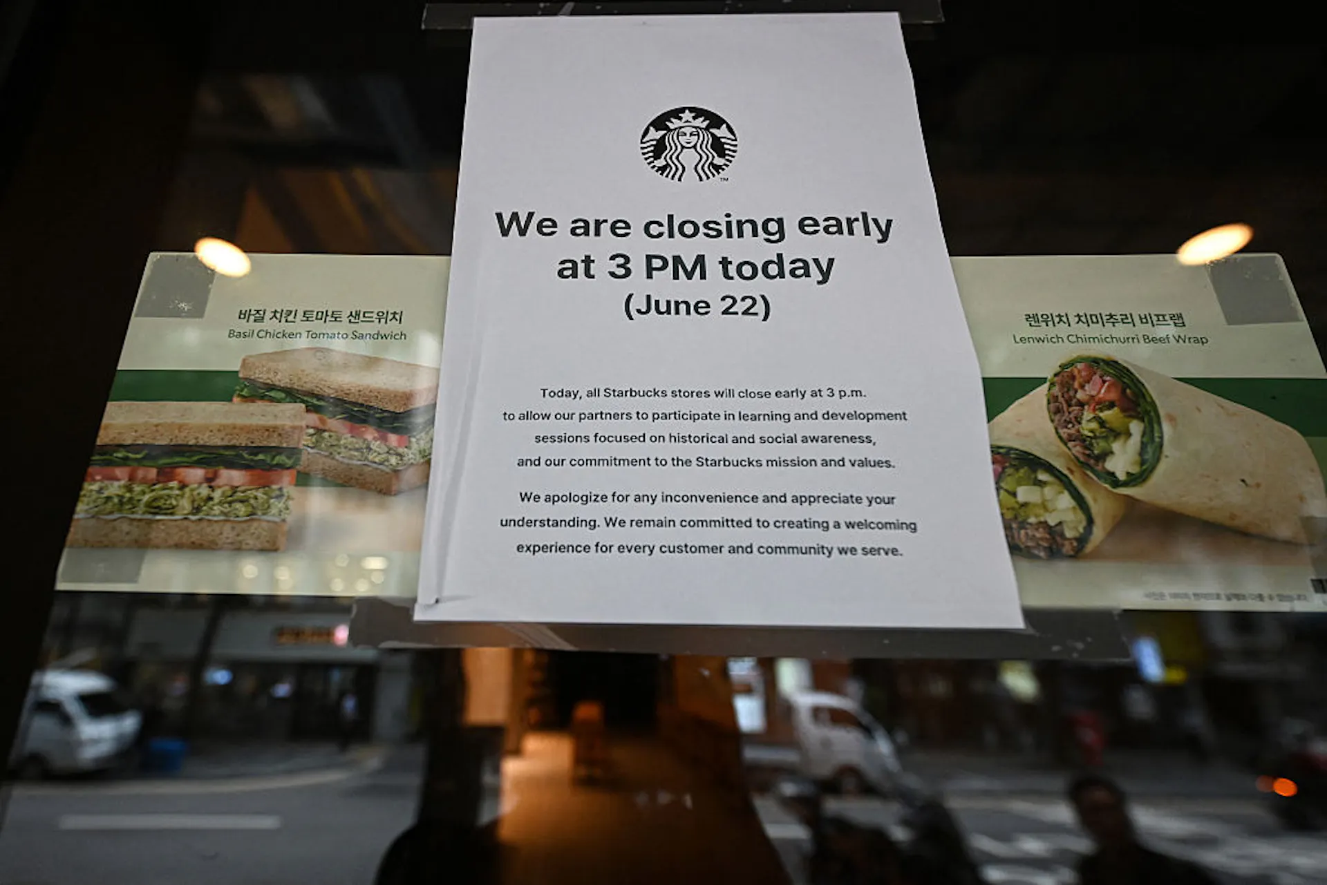 A glass window of a Starbucks location featuring posters of sandwiches and wraps alongside a white sign announcing an early closing so staff could undergo historical and social awareness training.
