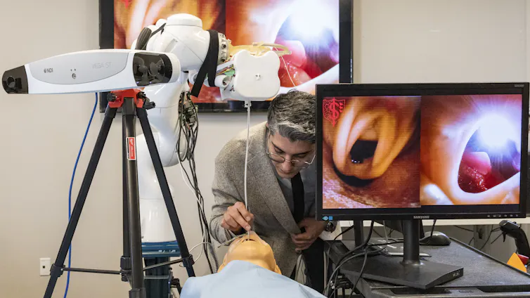 Un hombre con gafas opera a un paciente simulado entre un robot y una gran pantalla de computadora.