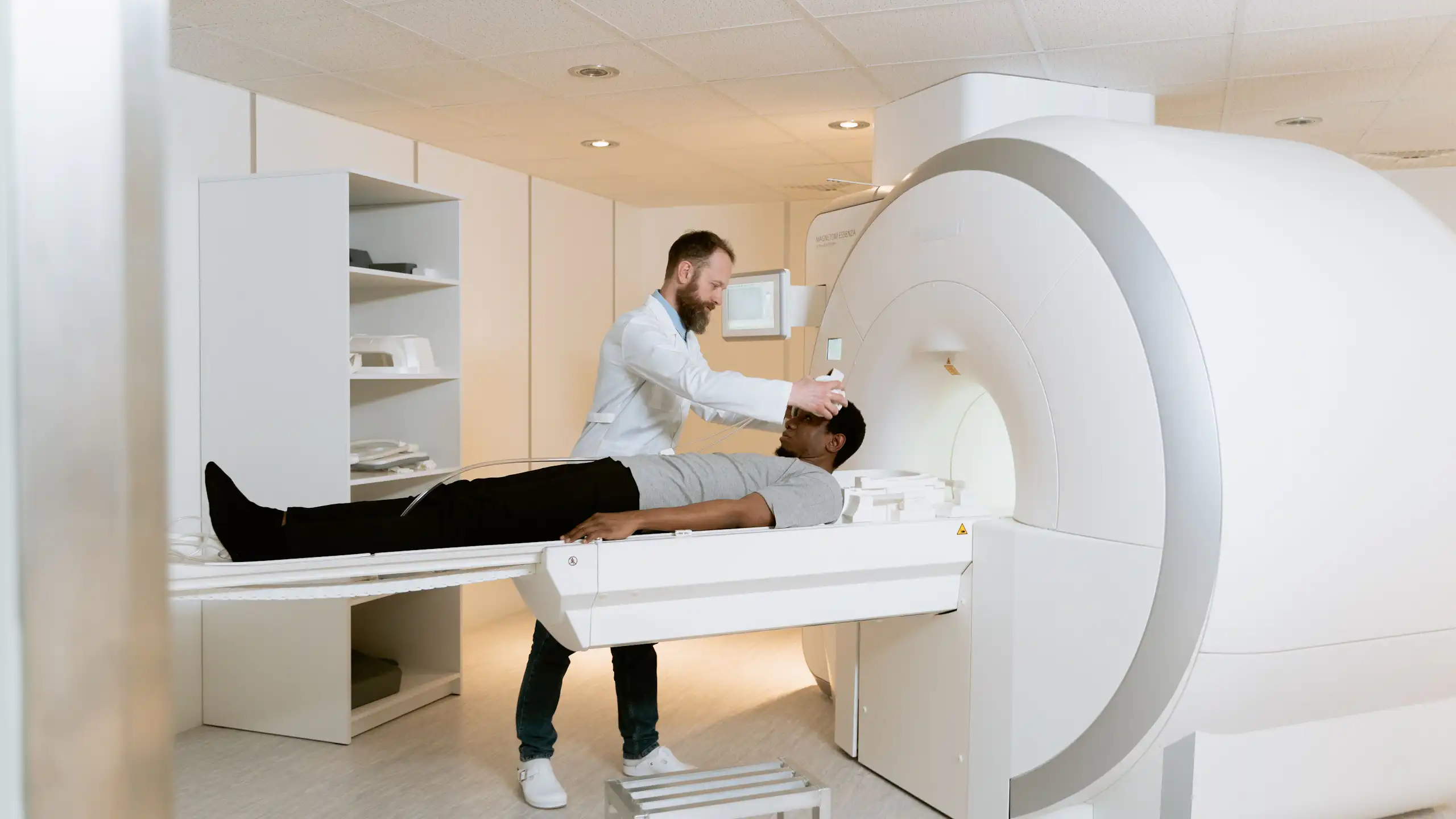 A Black man lies flat while a white man with a beard and a white coat prepares him to enter a diagnostic machine.
