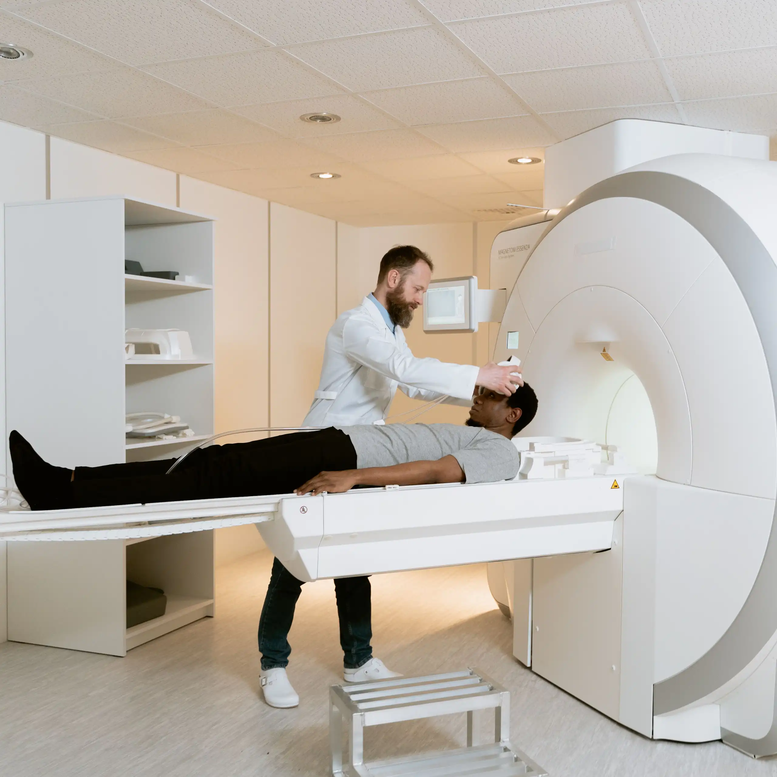 A Black man lies flat while a white man with a beard and a white coat prepares him to enter a diagnostic machine.
