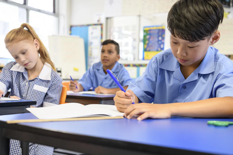 A young boy in a blue shirt with other pupils sitting in classroom doing their school work.
