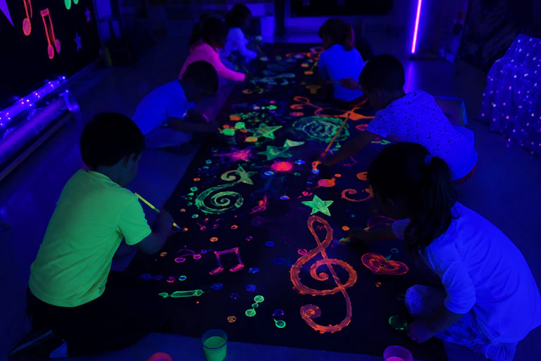 En una sala a oscuras, niños y niñas en cuclillas frente a un gran mural dibujan formas con pintura fluorescente que destacan en la oscuridad.