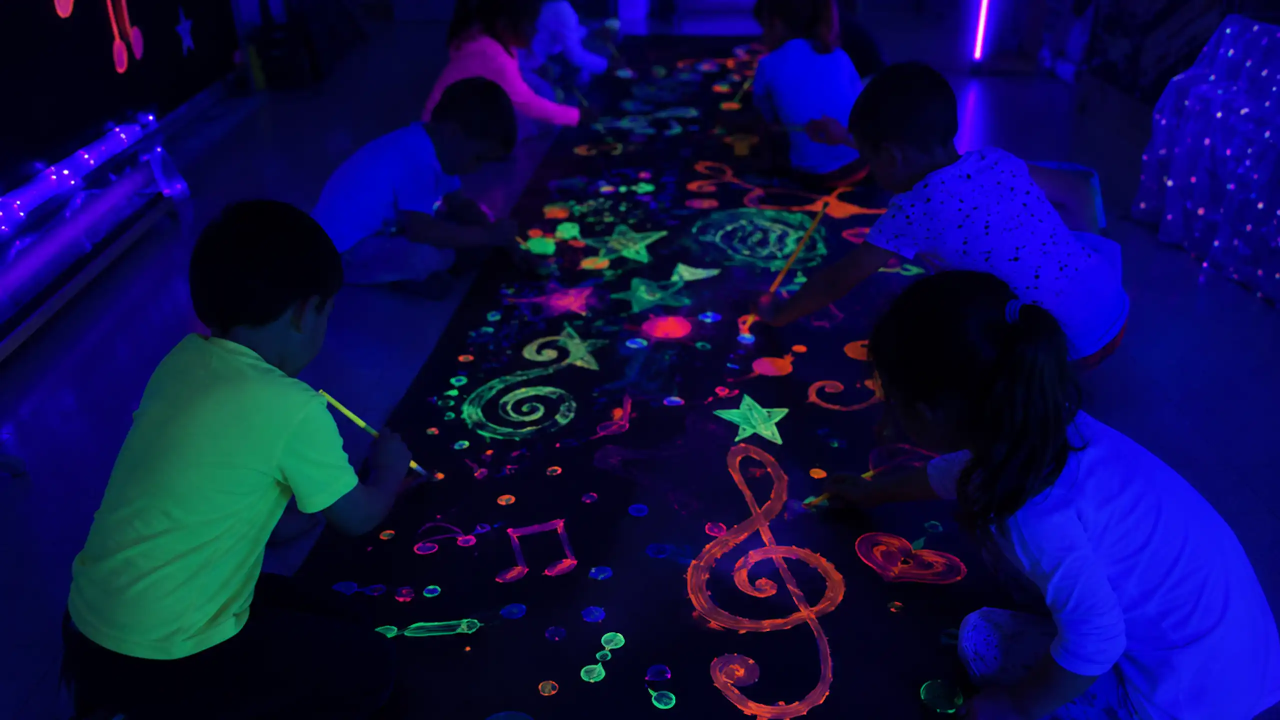 En una sala a oscuras, niños y niñas en cuclillas frente a un gran mural dibujan formas con pintura fluorescente que destacan en la oscuridad.