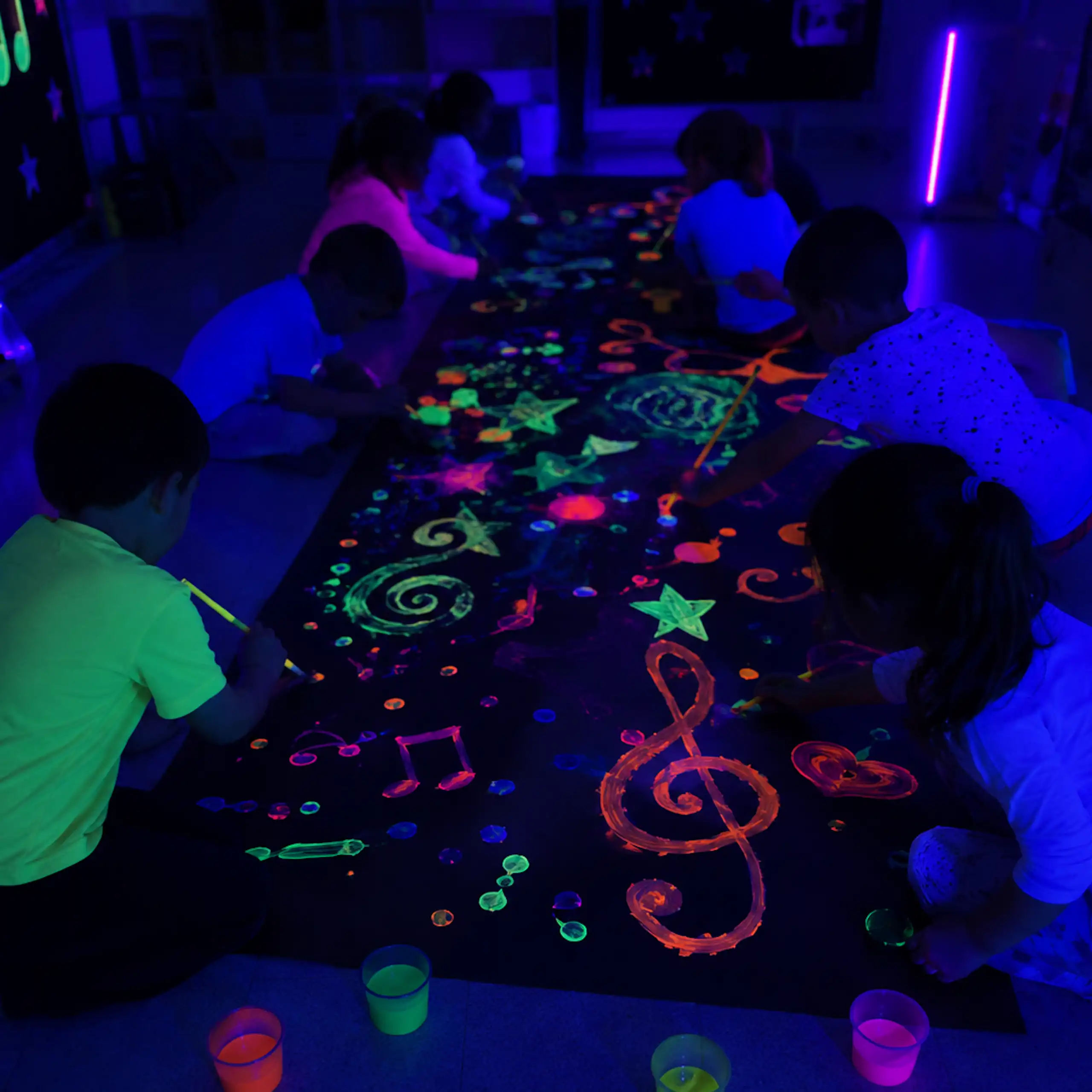 En una sala a oscuras, niños y niñas en cuclillas frente a un gran mural dibujan formas con pintura fluorescente que destacan en la oscuridad.