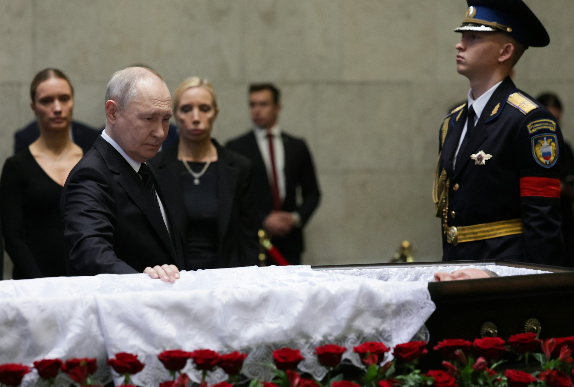 Russian president, Vladimir Putin, with his hand on a coffin while a soldier looks on.