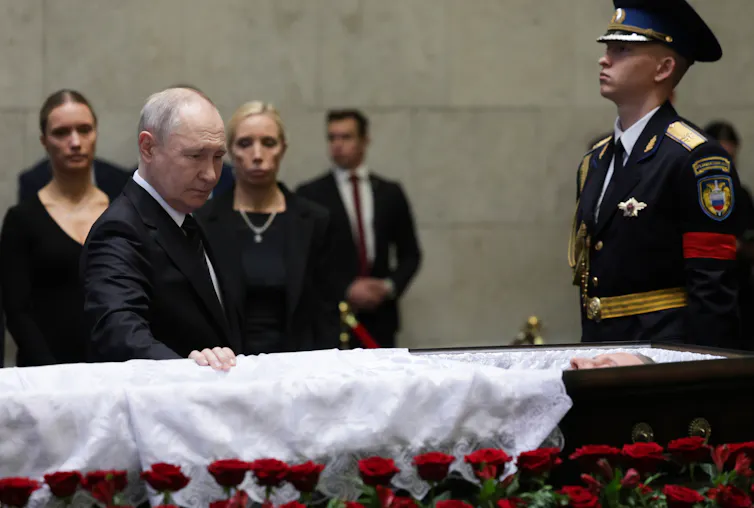 Russian president, Vladimir Putin, with his hand on a coffin while a soldier looks on.