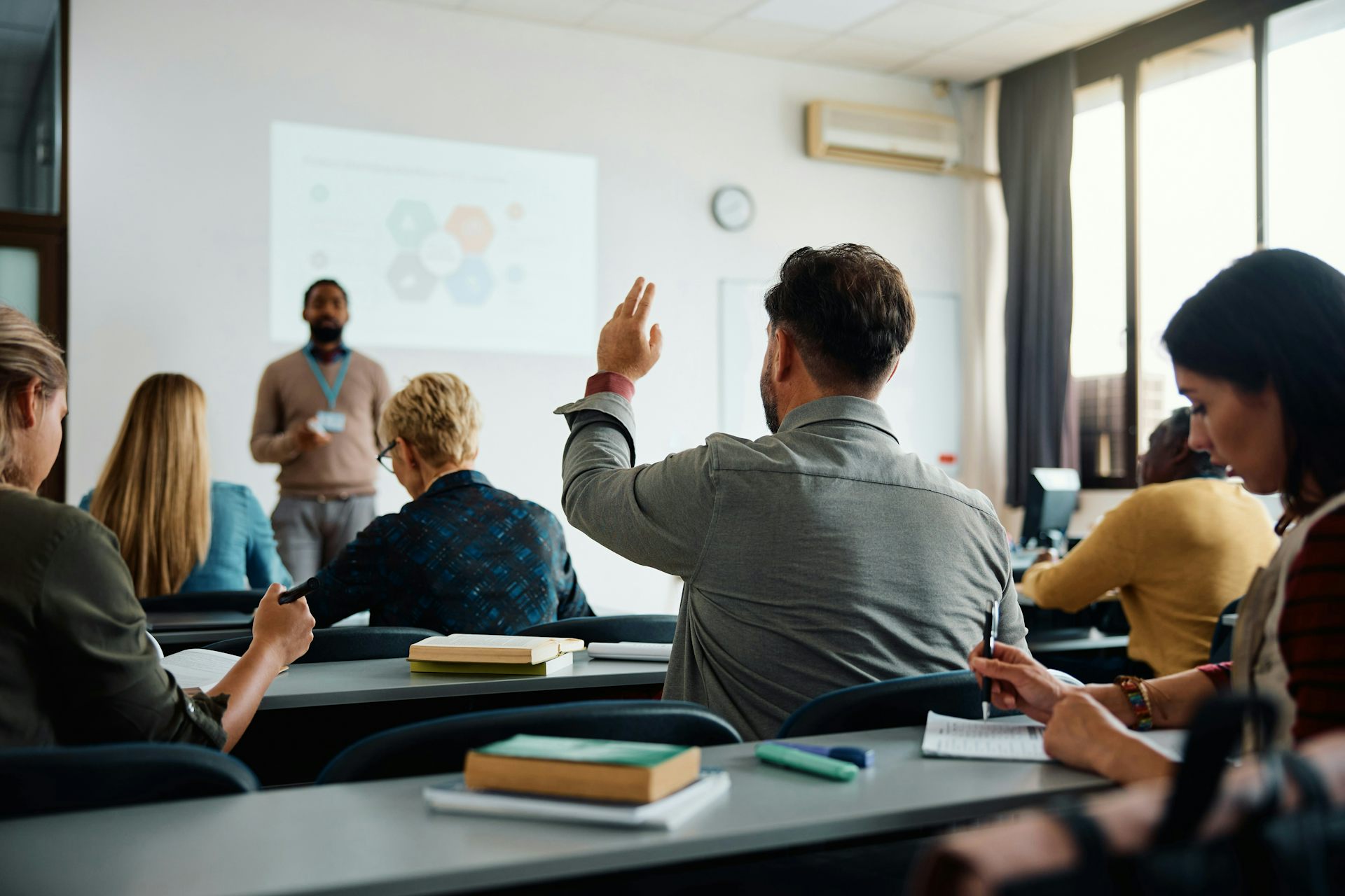 A small adult classroom where a young man has his hand raised an instructor is at the front listening.