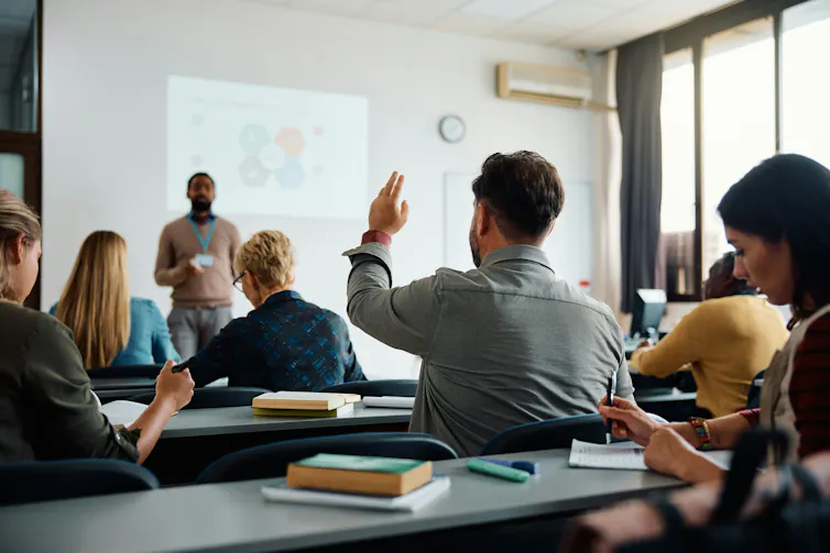 A small adult classroom where a young man has his hand raised an instructor is at the front listening.