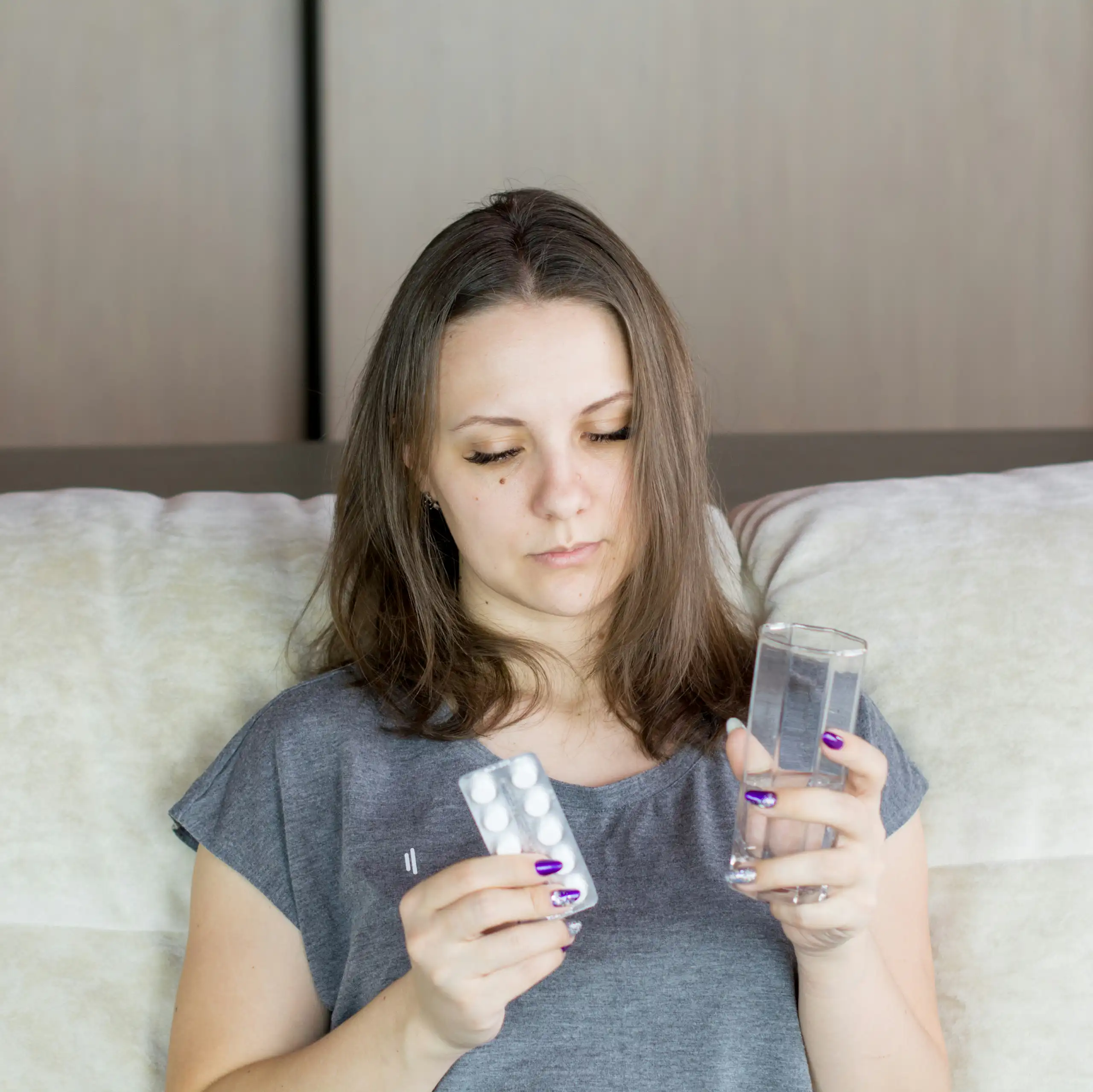 A woman sitting on a sofa looking at a blister pack of medication and holding a glass of water