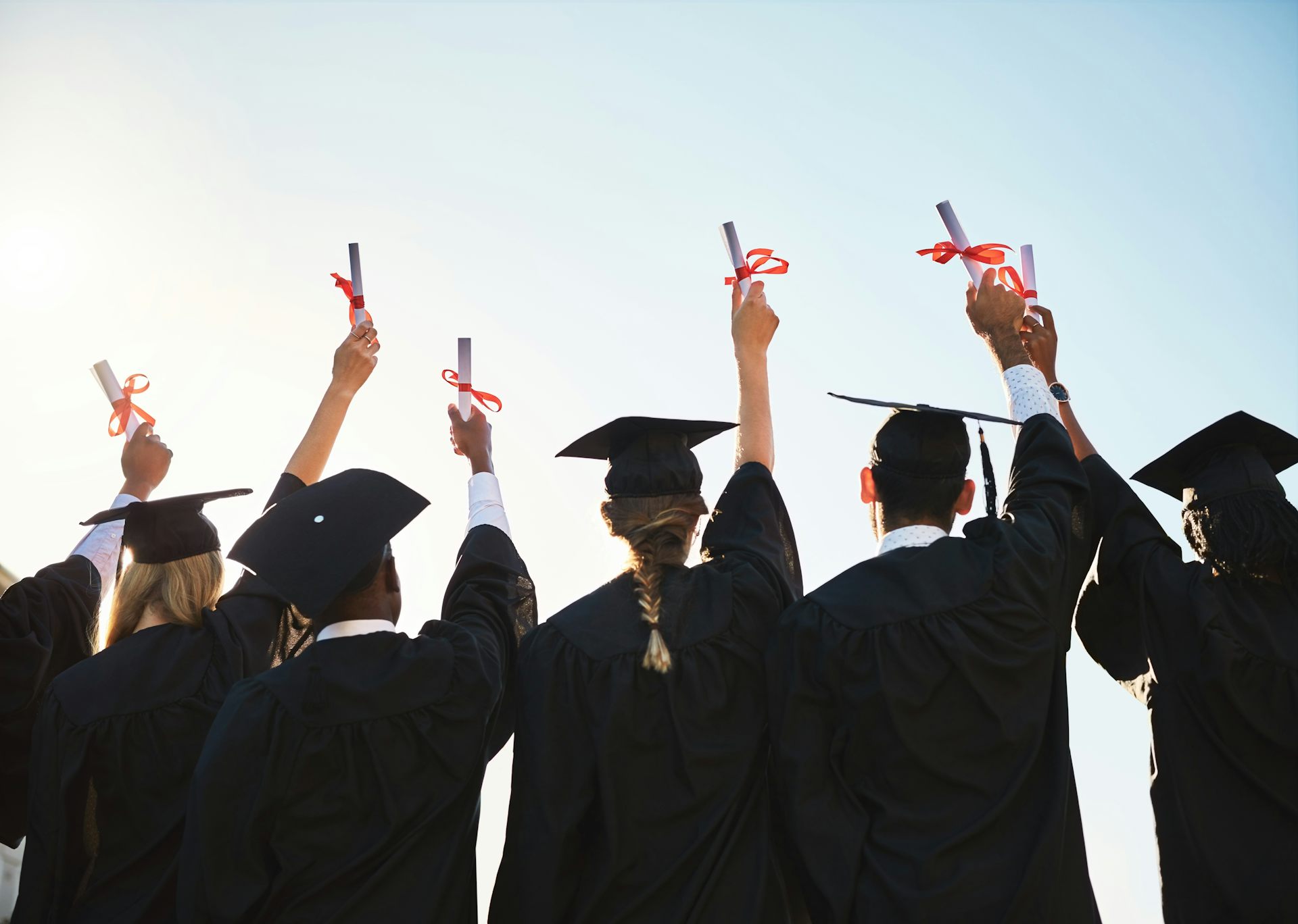 Estudiantes graduados de espaldas con birrete levantando el título enrollado envuelto con un lazo rojo