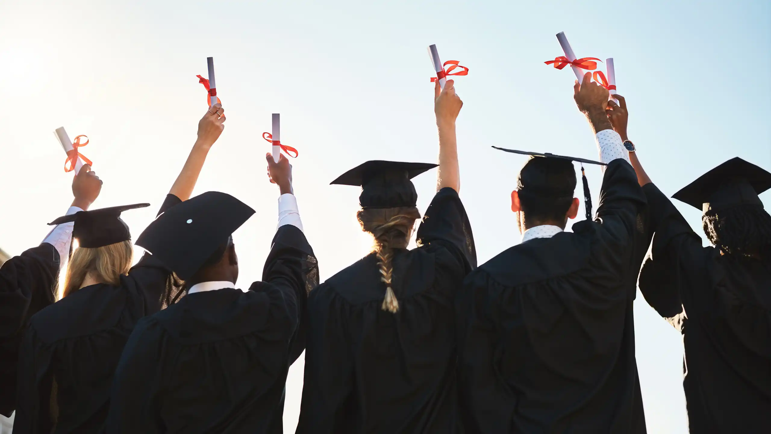 Estudiantes graduados de espaldas con birrete levantando el título enrollado envuelto con un lazo rojo