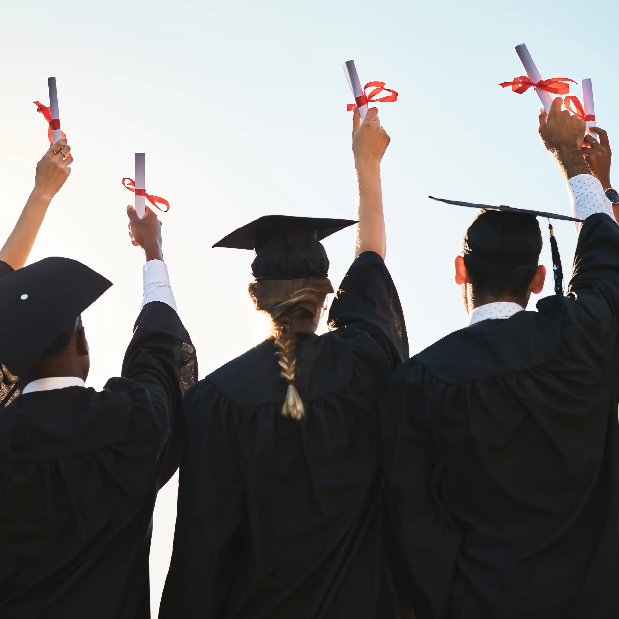 Estudiantes graduados de espaldas con birrete levantando el título enrollado envuelto con un lazo rojo