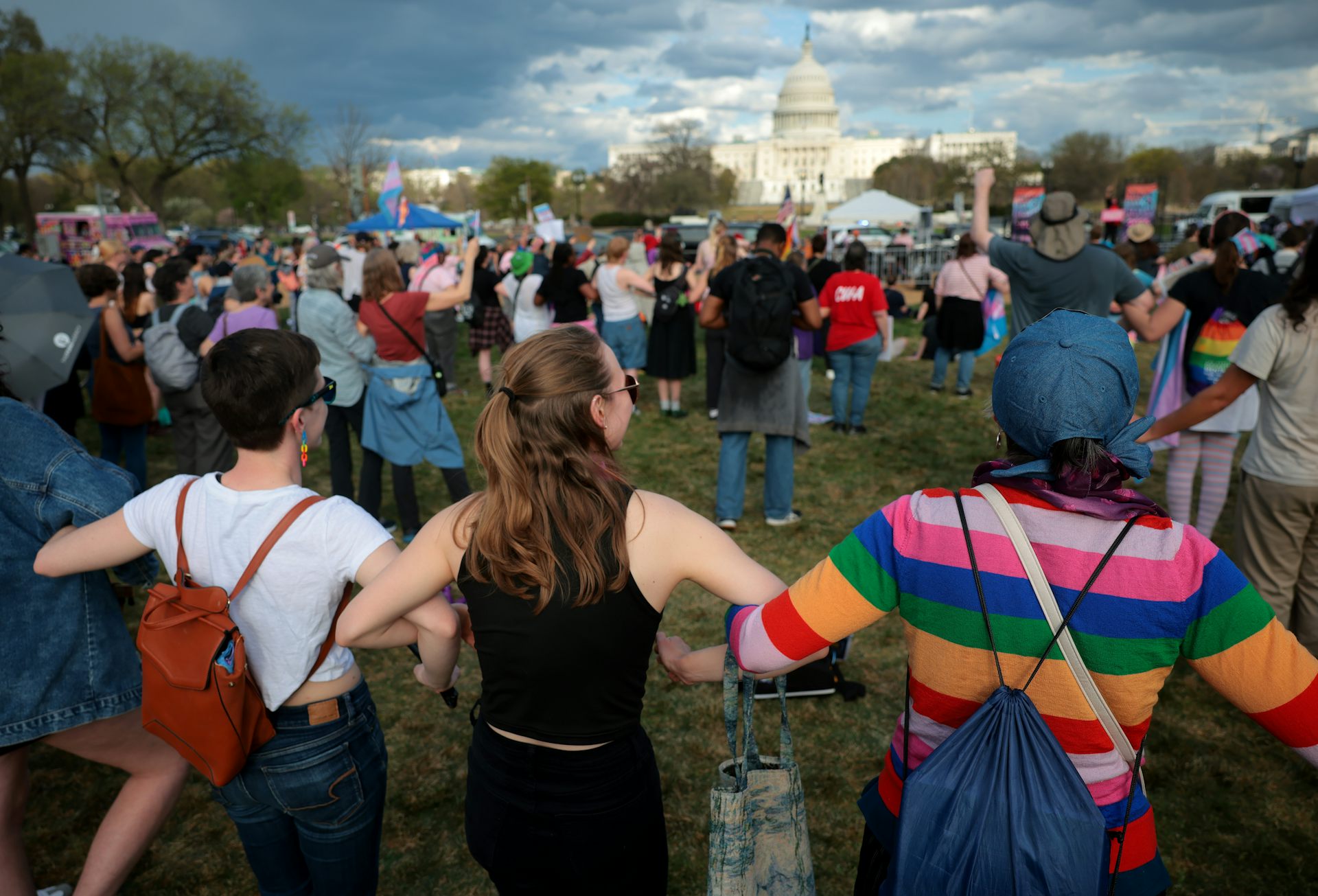 People stand with linked arms in a crowd facing the U.S. Capitol, transgender pride flags among them