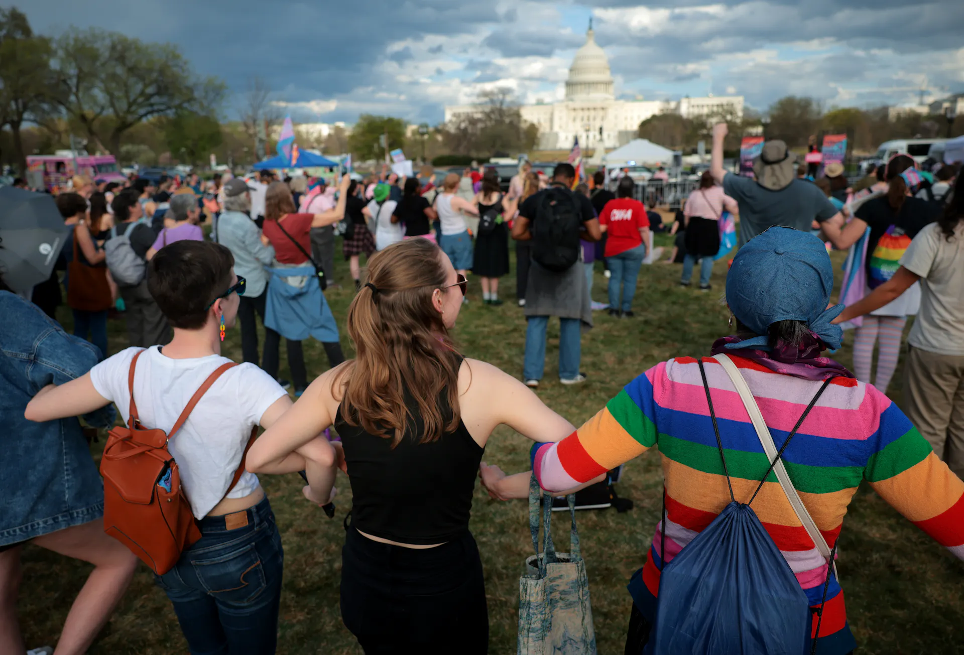 People stand with linked arms in a crowd facing the U.S. Capitol, transgender pride flags among them