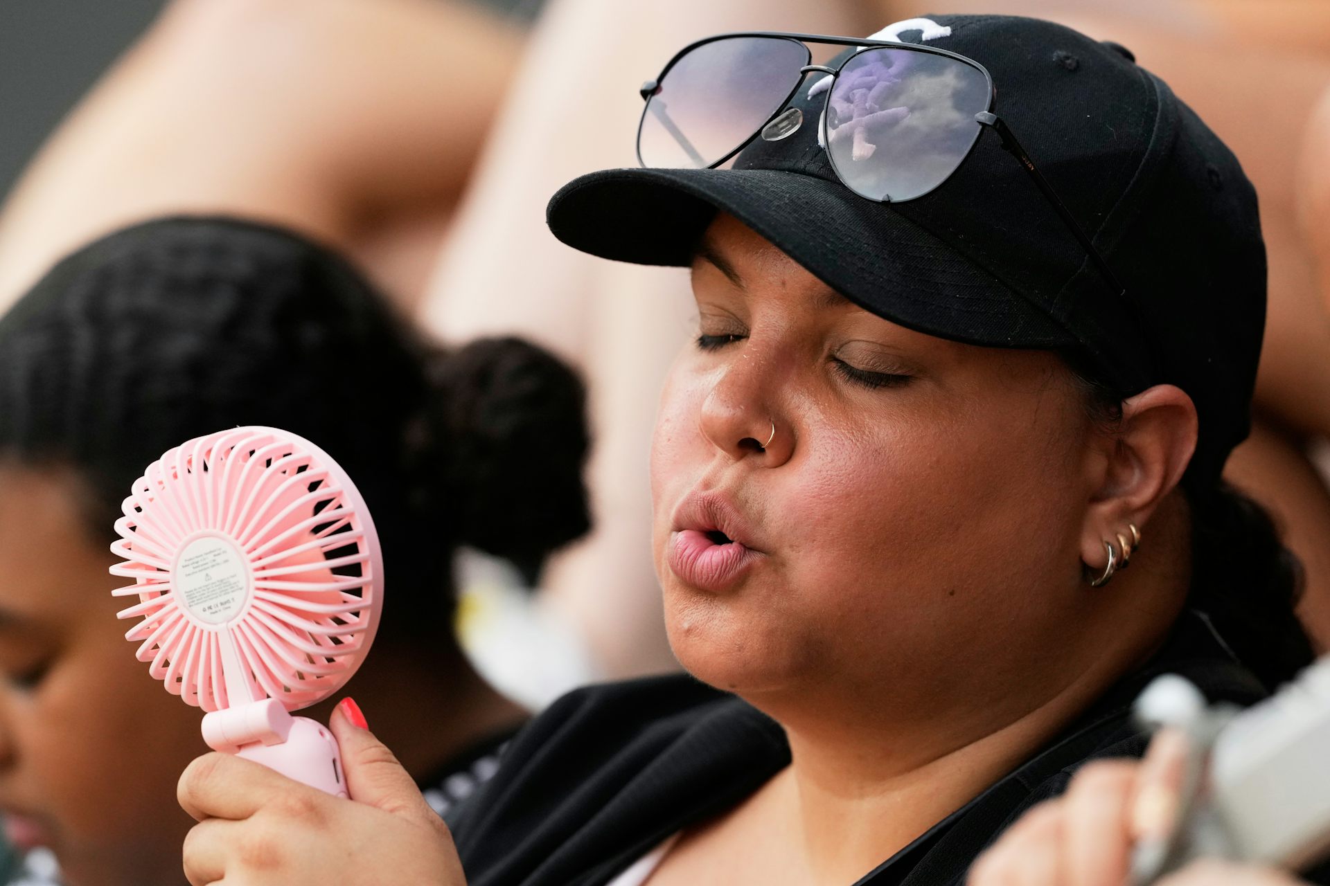 A woman outdoors in a baseball cap fans herself with a handheld fan