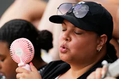 A woman outdoors in a baseball cap fans herself with a handheld fan