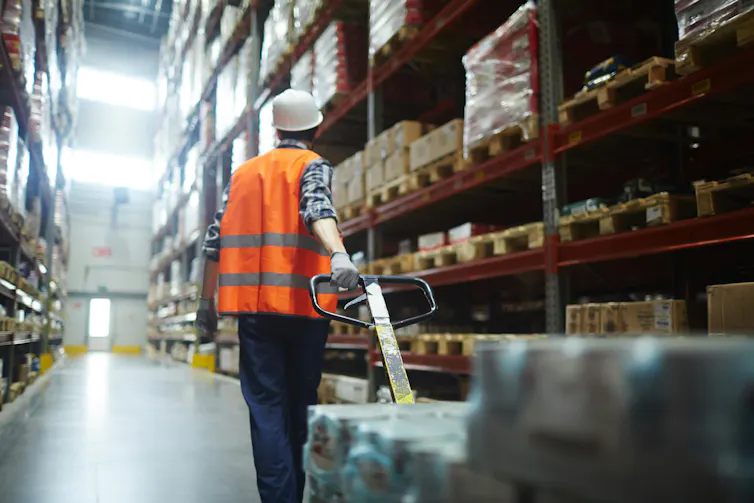 A worker in an orange vest pulls a trolley through a warehouse
