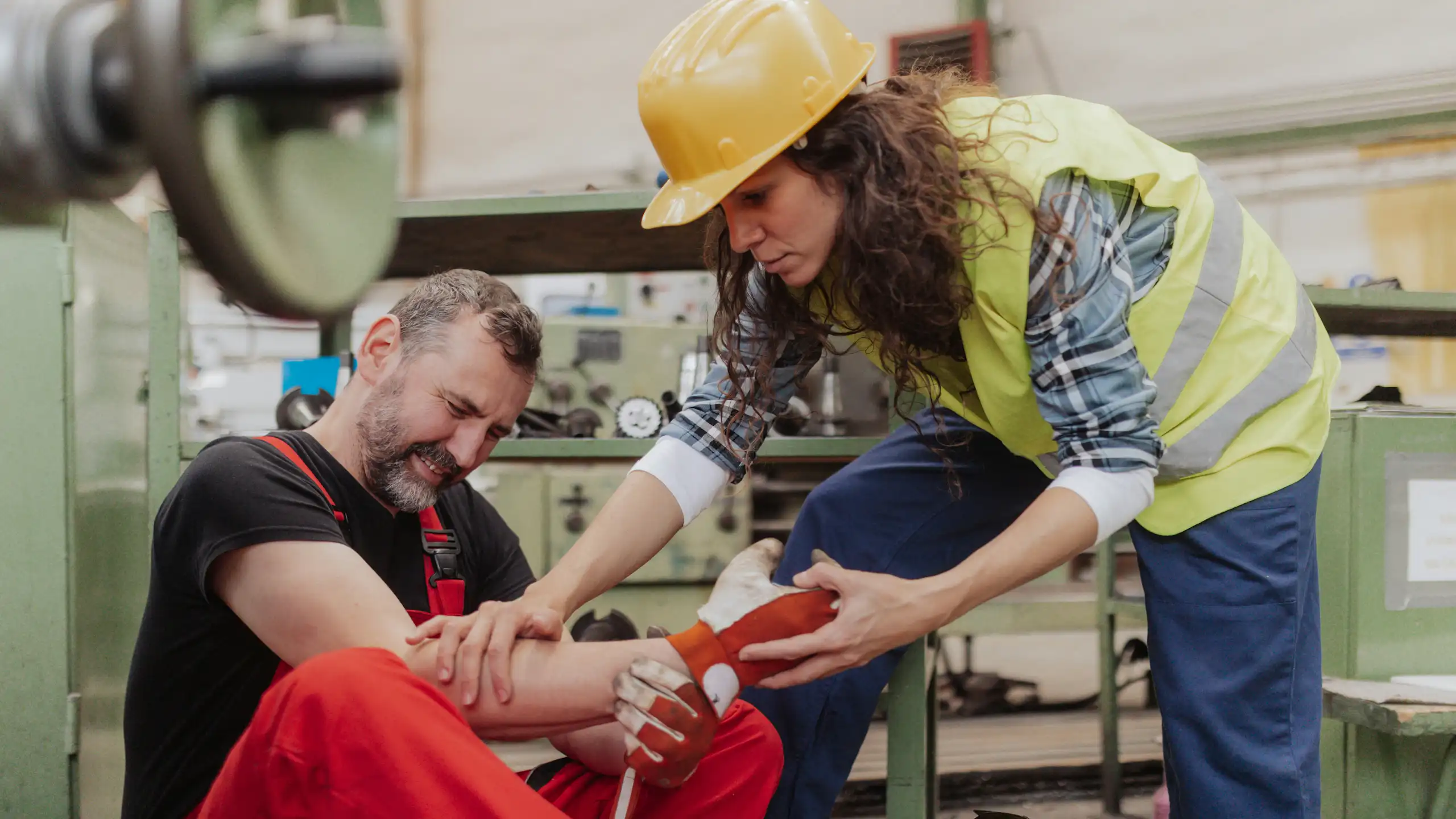 A woman in a hard hat offerss help to a man sitting on the ground, grimacing in pain