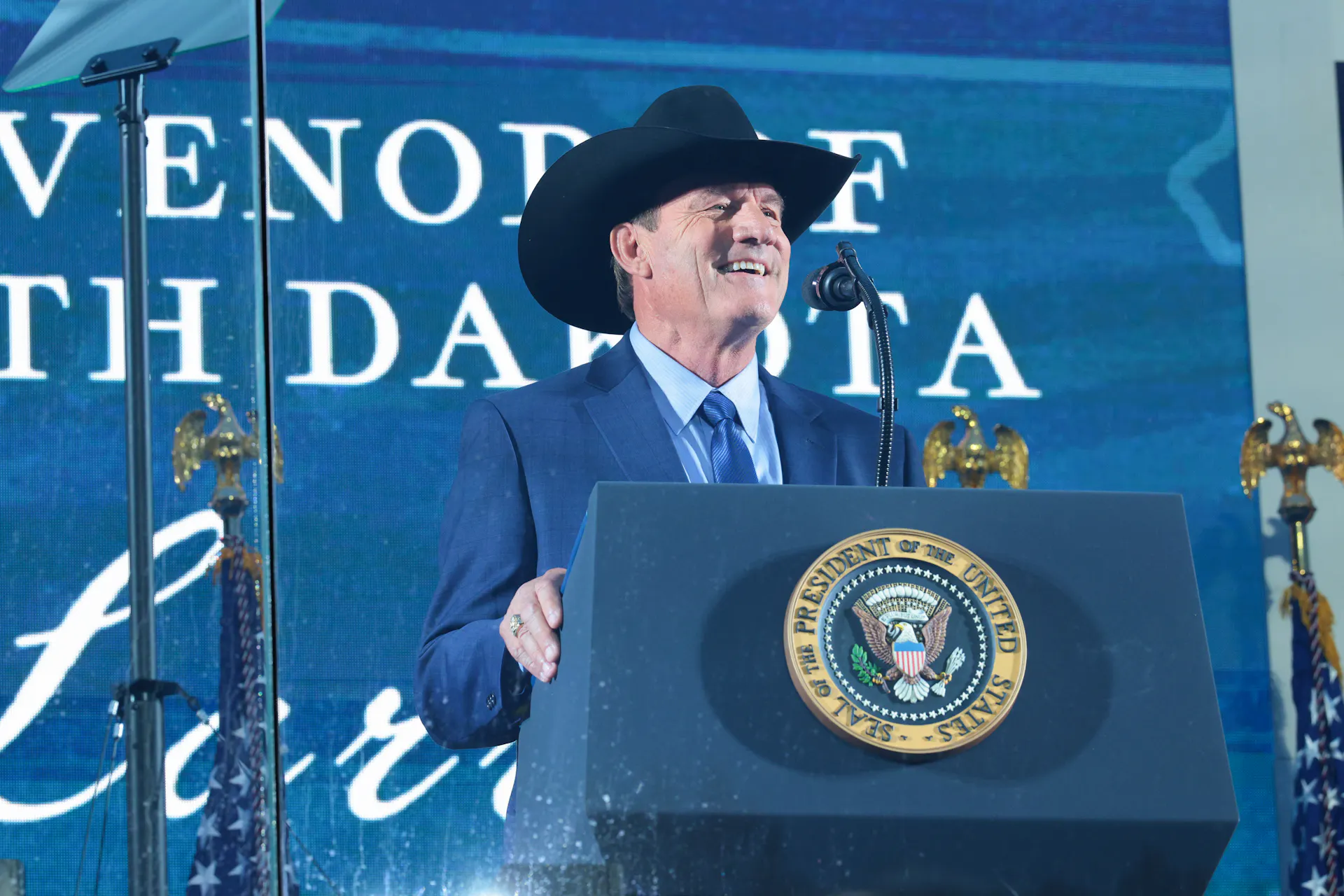 A man in a blue suit and wearing a cowboy hat gives a speech.