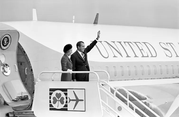 A man, waving, and a woman at the top of a stairway leading down from an airplane with 'UNITED STATES OF AMERICA' painted on its side.