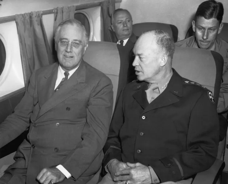 Two men, one in a suit and the other in a military uniform, sitting next to each other and talking on an airplane.