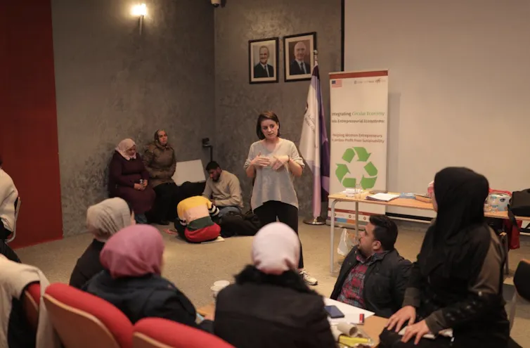 A group of students listen to an instructor, in a white shirt, standing next to a poster with a recycling logo.