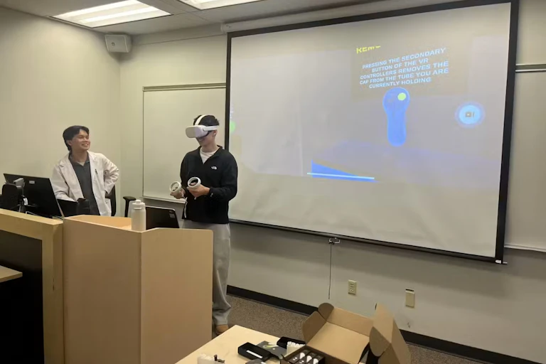 Two young men stand at a podium in a lecture theatre. One wears a white lab coat and the other wears a virtual reality headset.