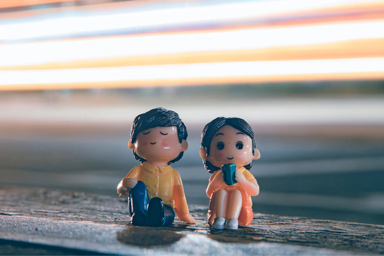 Two cute figurines sitting by a city roadside with vibrant light trails in the evening.