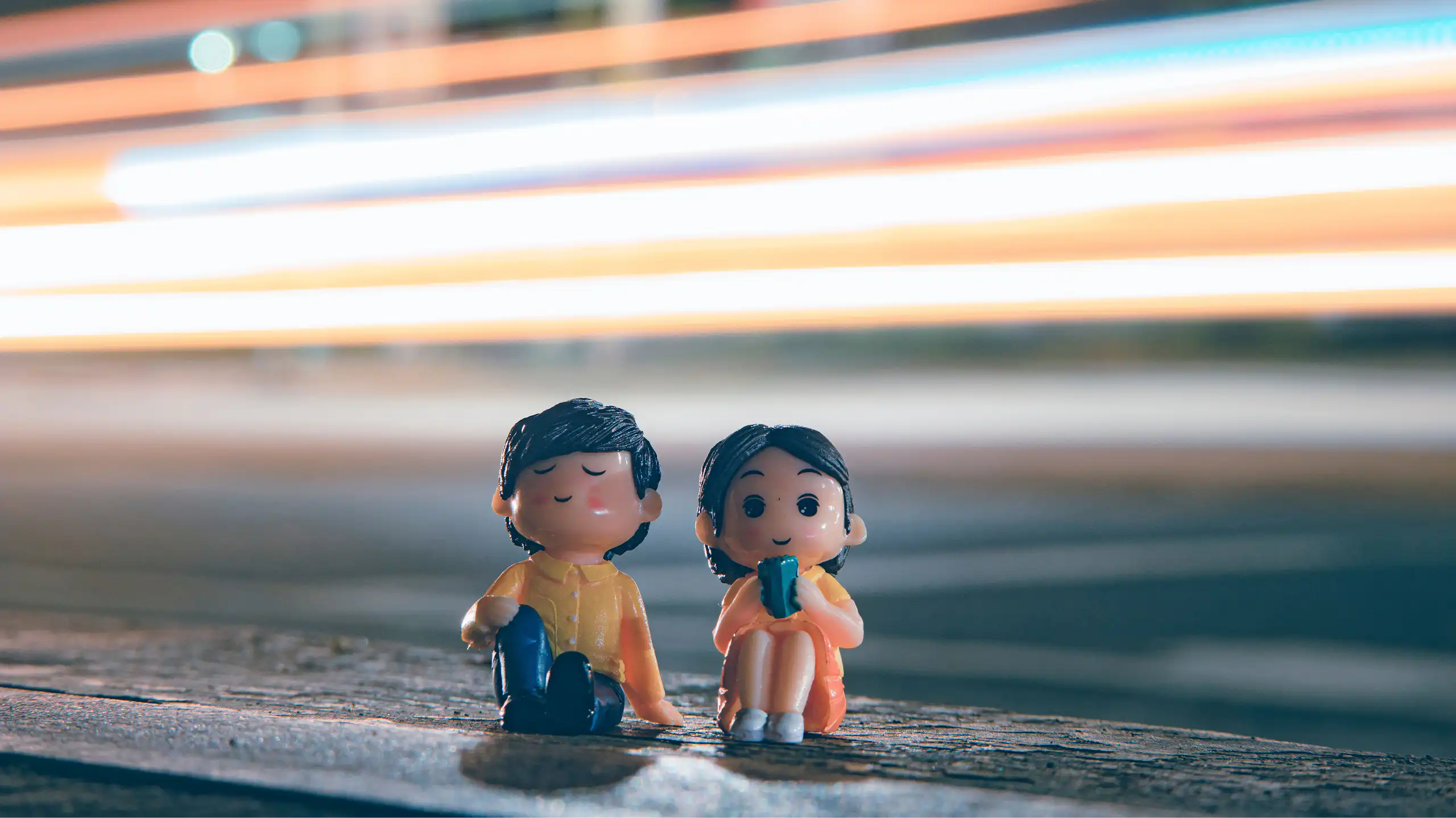 Two cute figurines sitting by a city roadside with vibrant light trails in the evening.