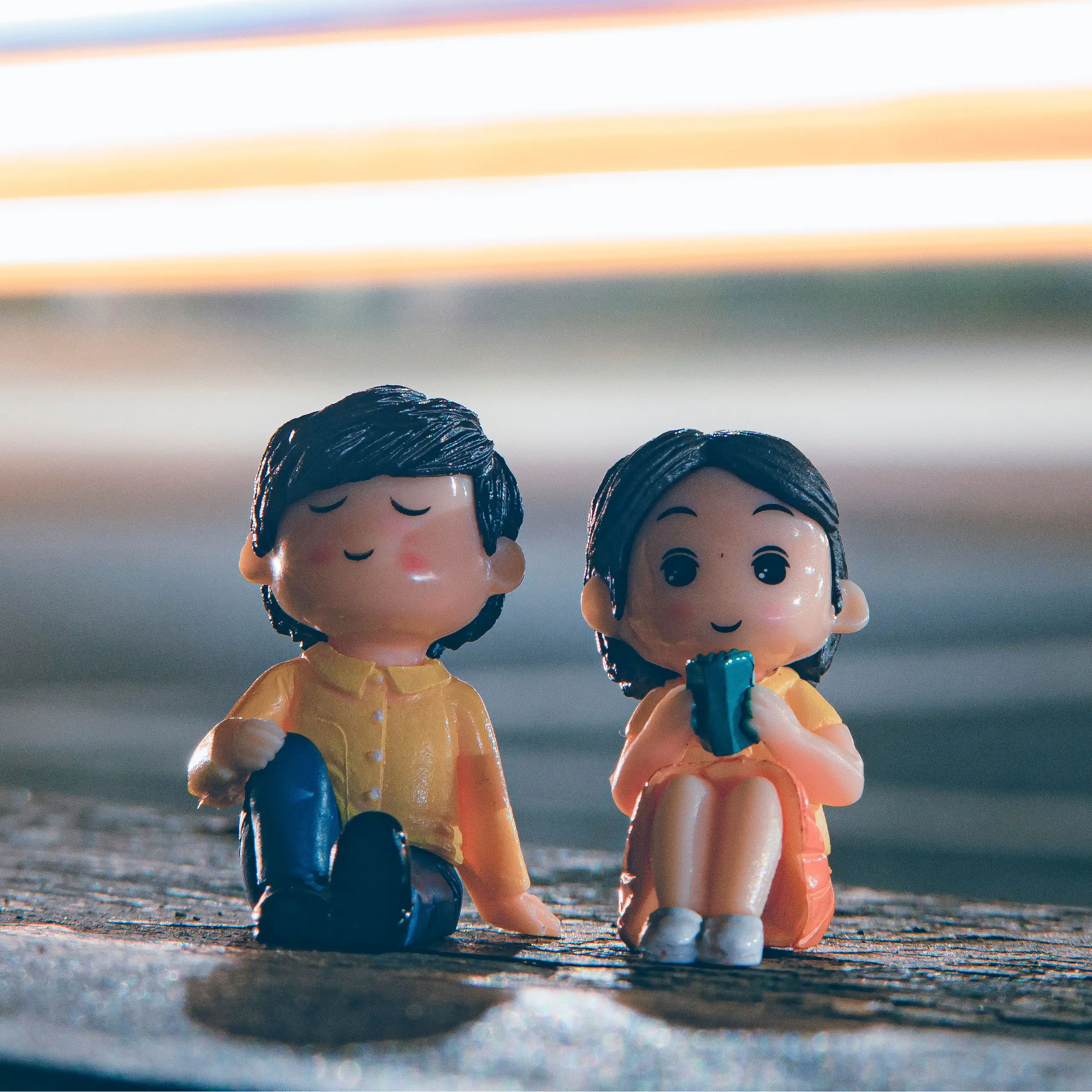 Two cute figurines sitting by a city roadside with vibrant light trails in the evening.