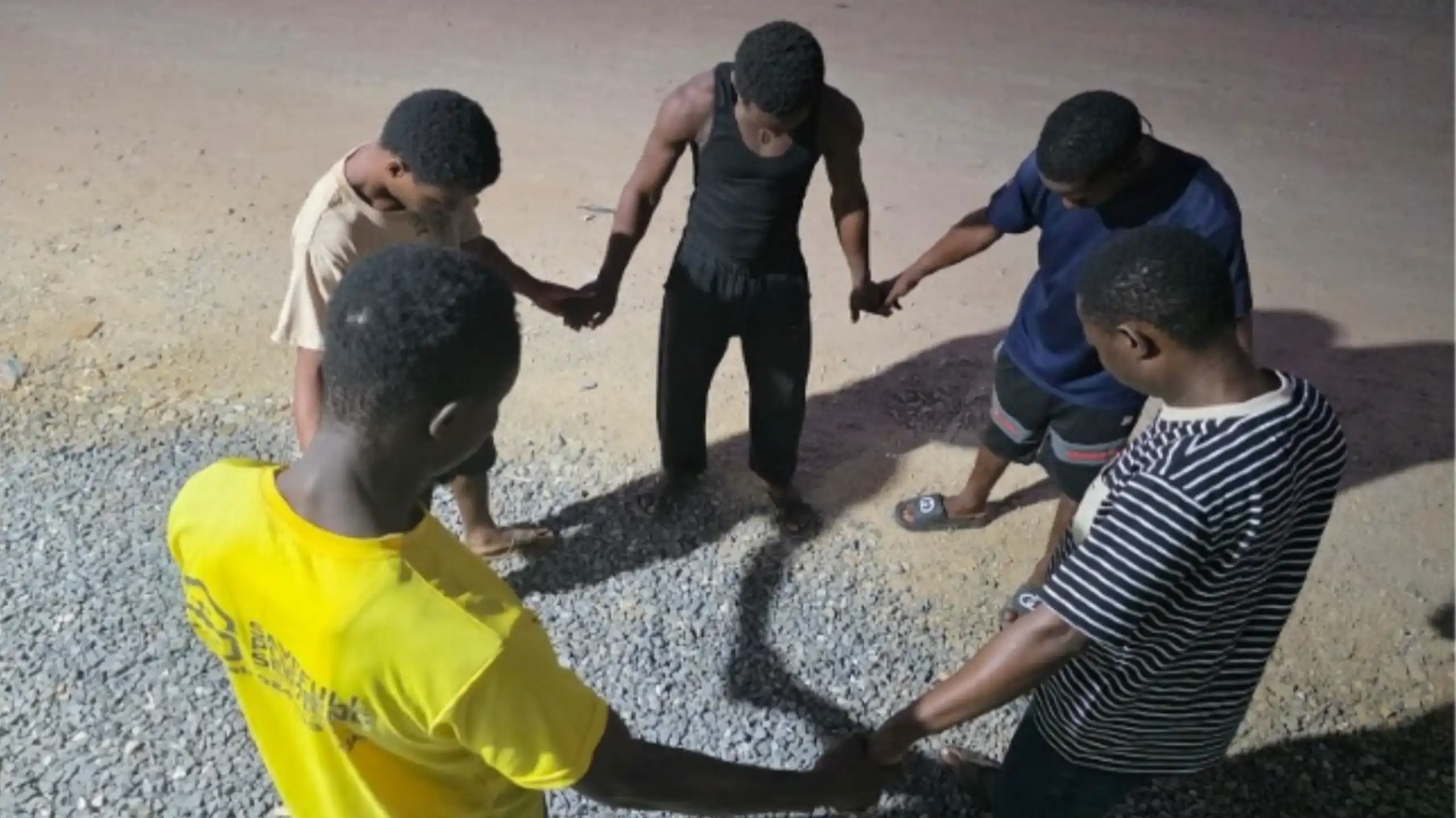 Young males stand in a circle, holding hands, with heads bowed