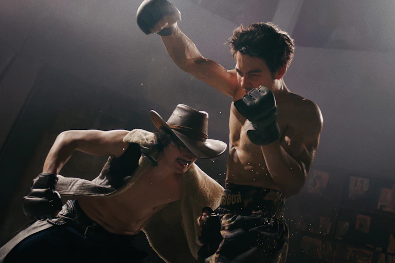 a shirtless boxer holds both hands up, looks like he is giving a punch: below him a man in a cowboy hat, vest and boxing gloves, ducks.