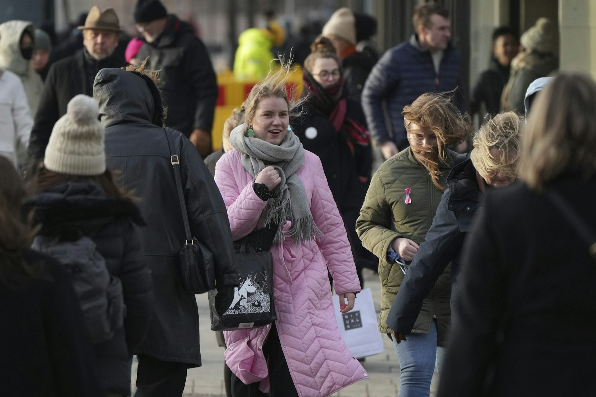 A smiling woman in a pink winter coat smiles as she walks along a city street.
