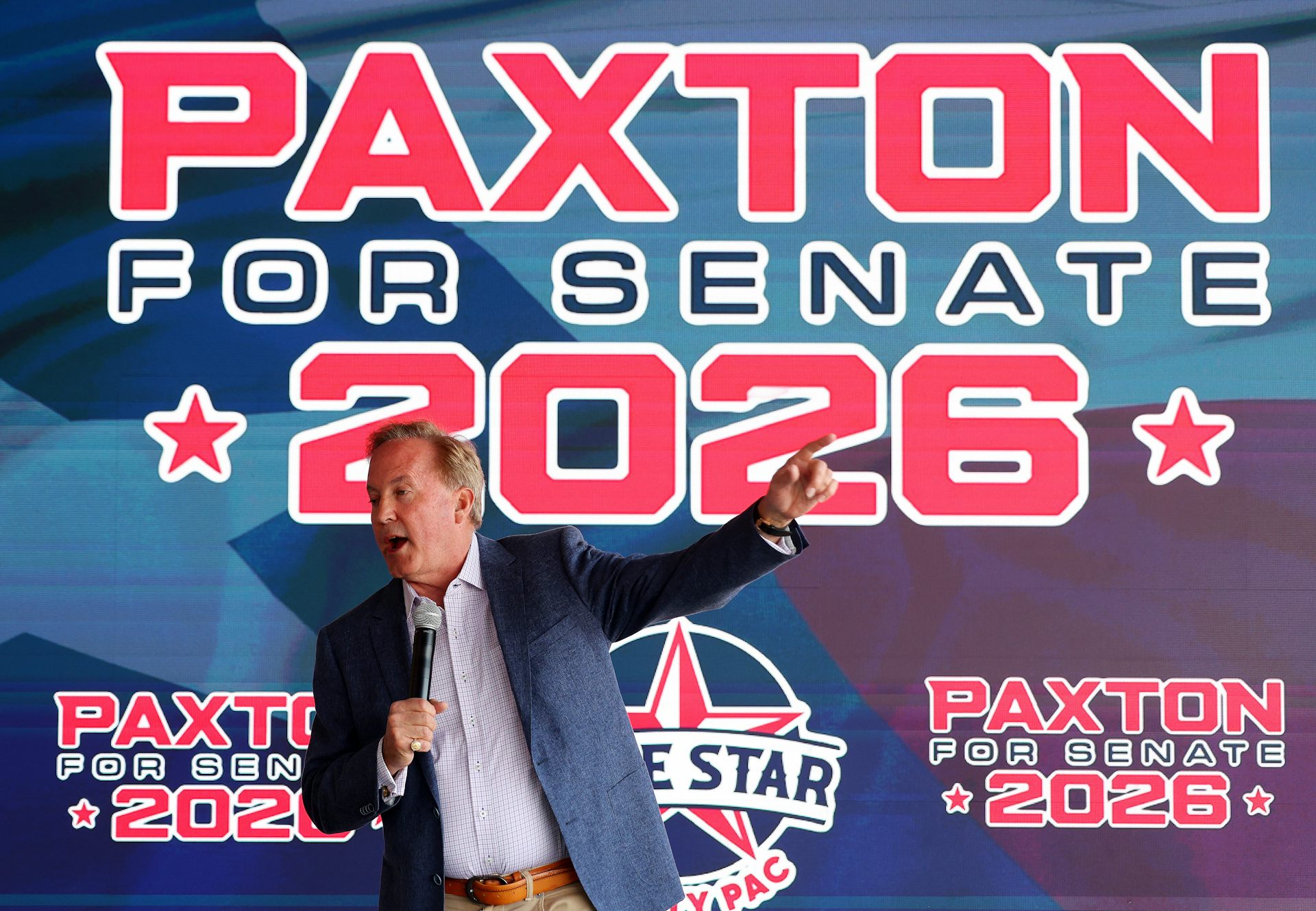 Middle-aged man wearing a suit jacket gestures and speaks into a microphone in front of a massive banner reading 'Paxton for Senate 2026.'