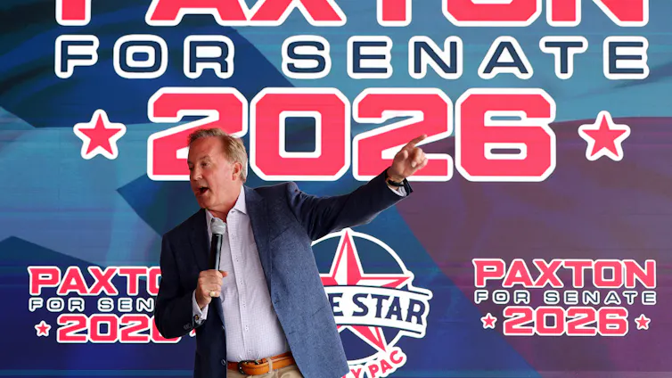 Middle-aged man wearing a suit jacket gestures and speaks into a microphone in front of a massive banner reading 'Paxton for Senate 2026.'