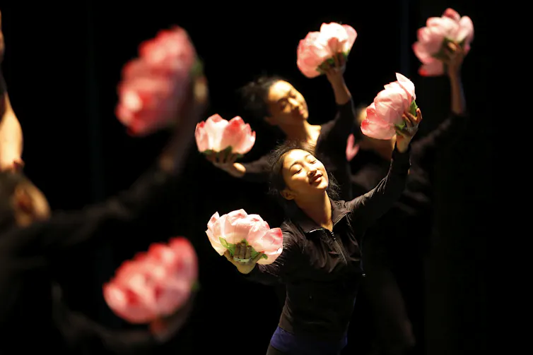 Chinese dancers with eyes closed hold large flowers during a performance.