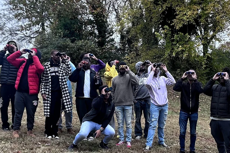 A group of college students lined up in a park, using binoculars to look up at the sky