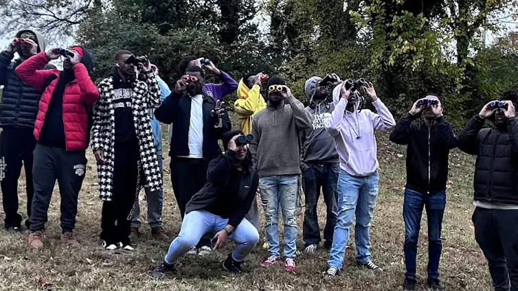 A group of college students lined up in a park, using binoculars to look up at the sky