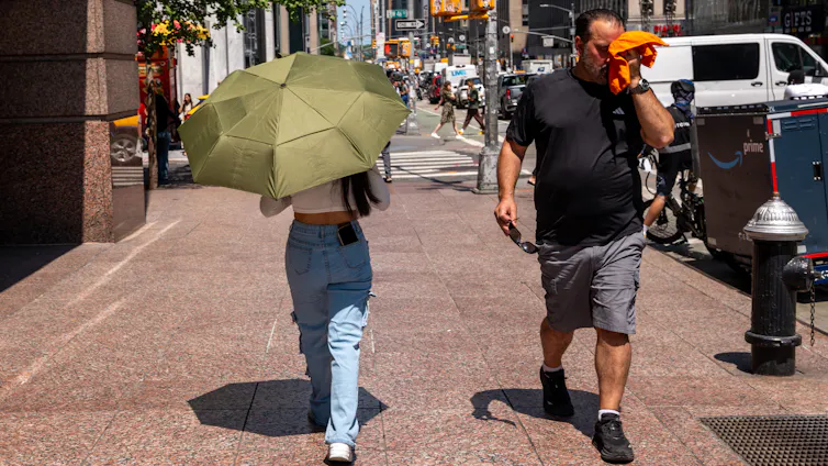 Two people walk in opposite directions on a city street, one carrying an umbrella for shade and the other wiping his face with a handkerchief.