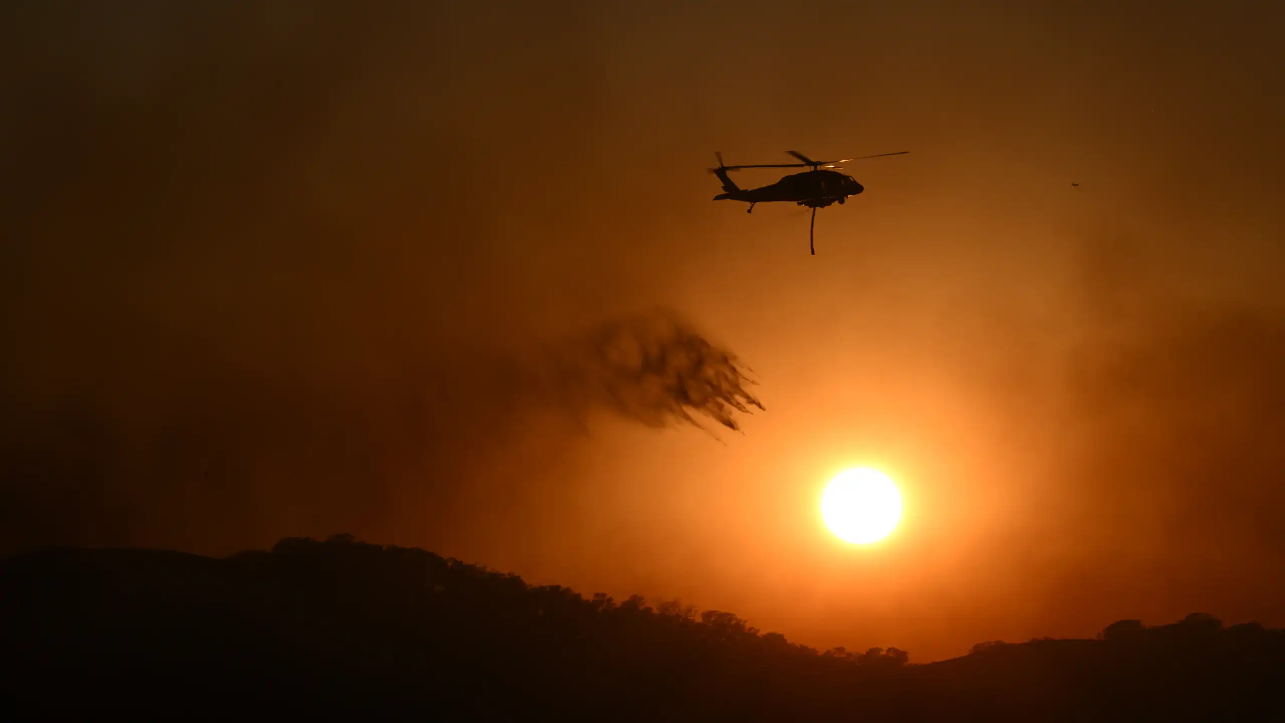 A fire helicopter drops water on a blaze in a dark sky.
