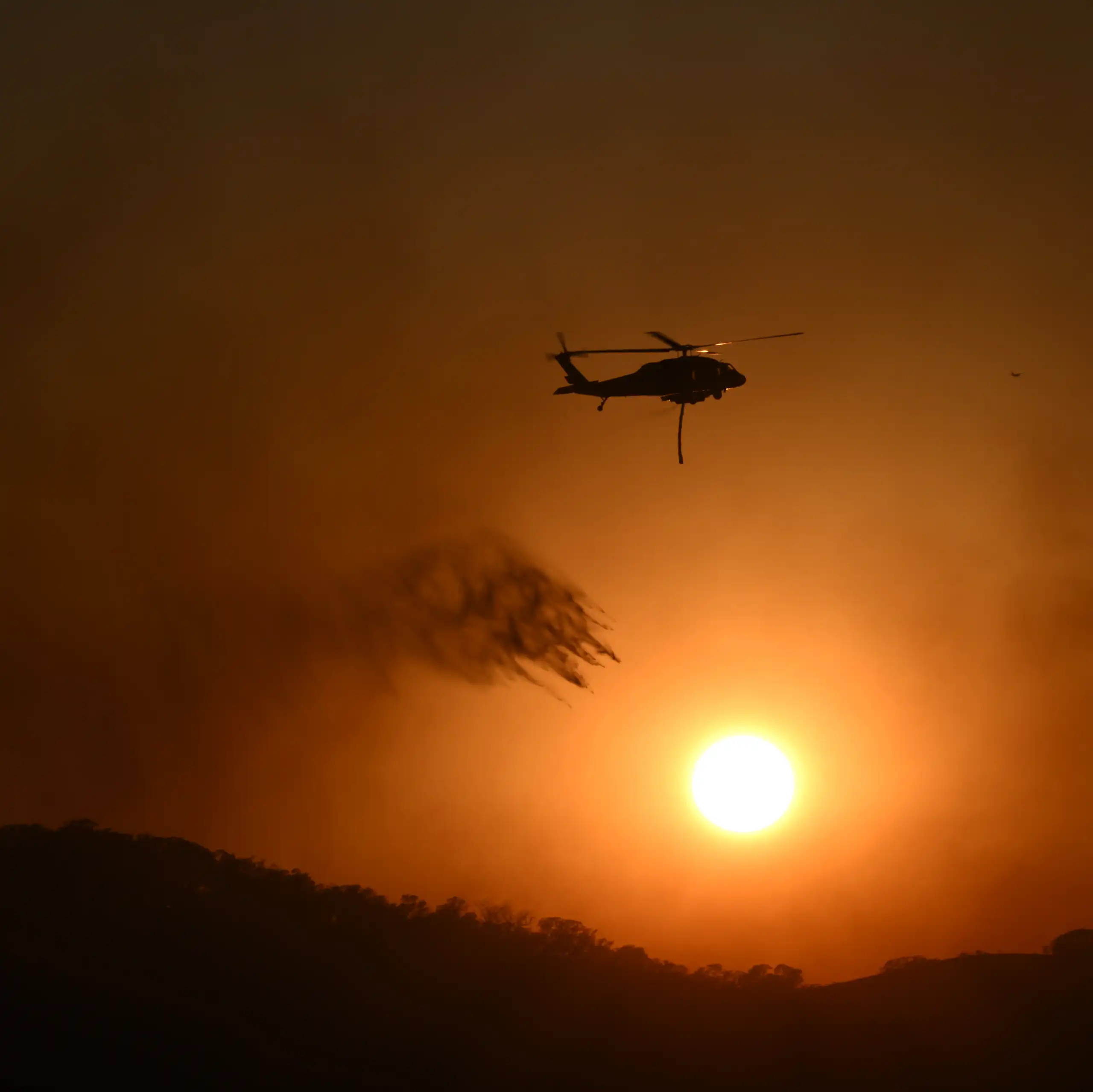 A fire helicopter drops water on a blaze in a dark sky.