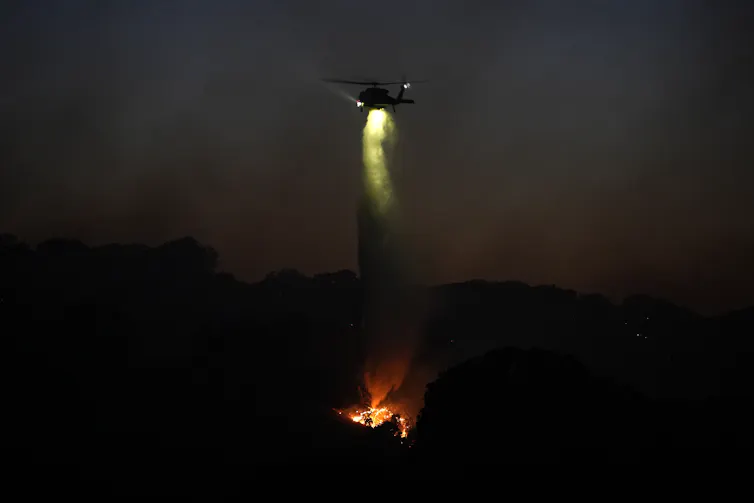 A fire helicopter drops water over flames in the dark as crews battle a wildfire.