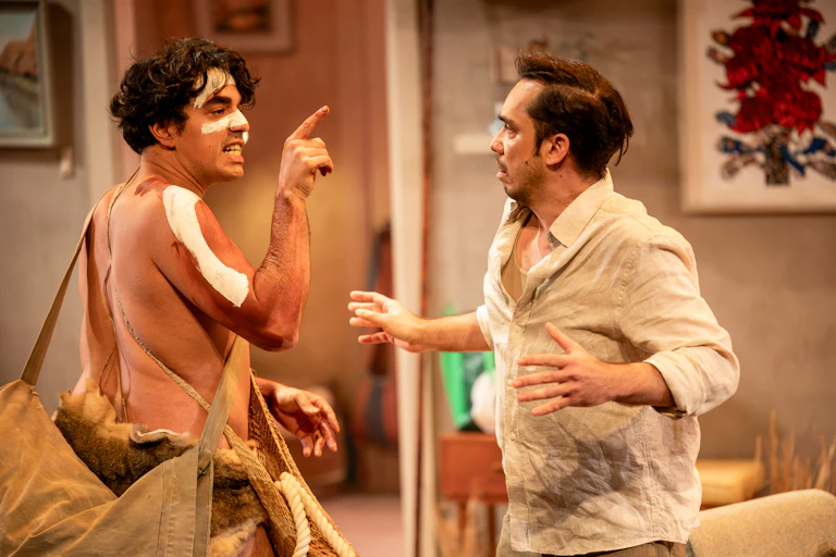 Two performers interact onstage in domestic set under warm lighting. One points his finger at the other.